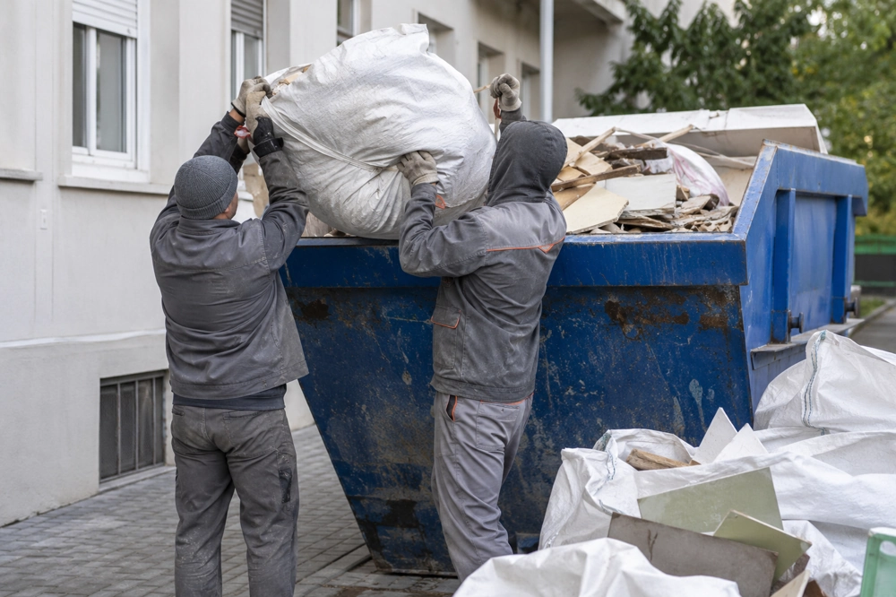 Dwóch pracowników przygotowuje wywóz odpadów budowlanych, wkładając worki do kontenera
